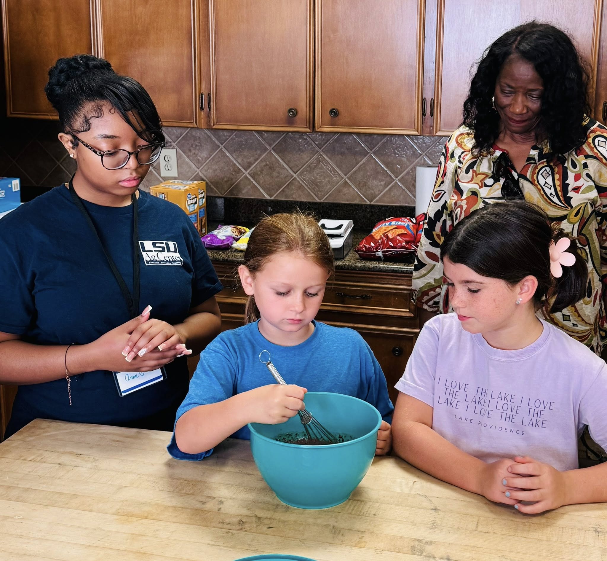 Two girls and a woman watching another girl whisk in a bowl.